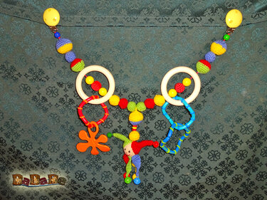 Gehäkelte Kinderwagenkette mit bunten Kugeln, Holzringen, Beißringen und einer kleinen Clownfigur, an Clips befestigt.