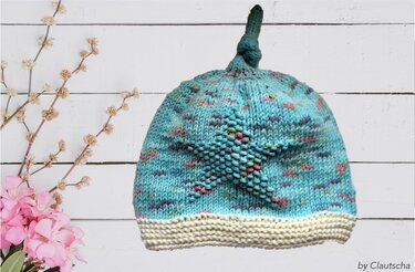 Türkise gestrickte Babymütze mit Sternmuster, heller Krempe und Knoten auf weißem Holzuntergrund