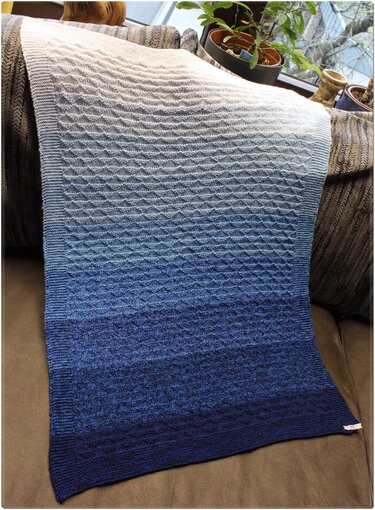 Gestrickte Babydecke mit Farbverlauf von Weiß zu Dunkelblau und strukturiertem Muster, auf einer Sofalehne drapiert.
