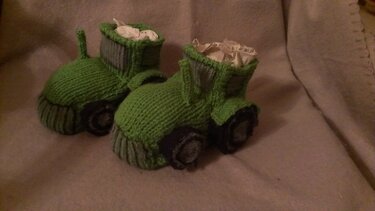 Zwei grüne, gestrickte Babyschuhe im Traktor‑Design mit Reifenapplikationen