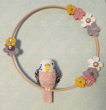 Gehäkelter rosa-weißer Amigurumi-Vogel auf Holz-Stickrahmen mit Häkelblumen