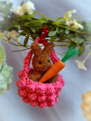 Gehäkelter brauner Hase mit Karotte in rosafarbenem Körbchen, hängend an einem Zweig