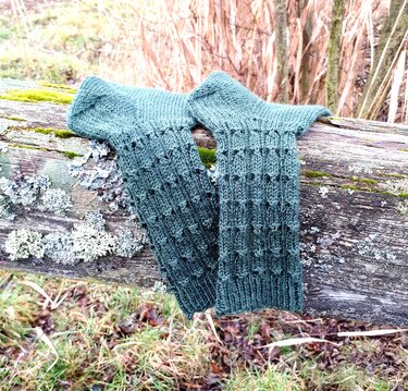 Zwei dunkelgrüne gestrickte Beinstulpen mit strukturiertem Muster auf einem moosbewachsenen Holzstamm.