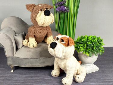 Two crocheted bulldog amigurumi toys: a brown-and-cream one on a small grey sofa and a cream-and-brown one on the floor beside plants.