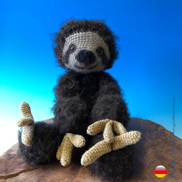 Gehäkeltes Amigurumi‑Faultier mit dunklem, flauschigem Körper und beigefarbenen Krallen auf einer Holzplatte vor blauem Himmel.