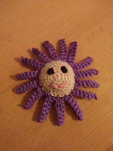 Handgehäkelte Sonne mit cremefarbenem Gesicht, schwarzen Augen, rosa Mund und lila spiralförmigen Strahlen