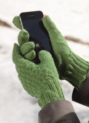 Hände in grünen gestrickten konvertierbaren Handschuhen bedienen ein Smartphone vor verschneiter Kulisse.
