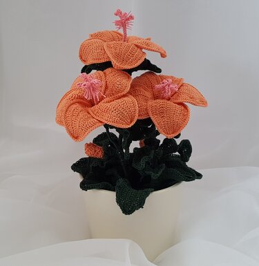 Gehäkelte Topfpflanze mit großen orangefarbenen Blüten, rosa Blütenstempeln und dunkelgrünen Blättern in hellem Topf vor hellgrauem Hintergrund.