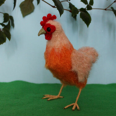 Gehäkeltes Amigurumi-Huhn in Orange und Pfirsich mit rotem Kamm auf grünem Stoff.