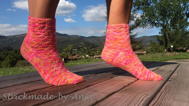 Kurzgestrickte Damensocken in Pink, Orange und Gelb mit Strukturmuster auf Holzterrasse vor Bergkulisse