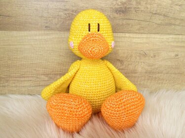 Gelbes gehäkeltes Amigurumi-Entenküken mit orangefarbenem Schnabel und Füßen auf weißem, flauschigem Untergrund.
