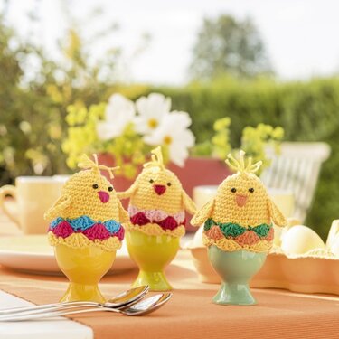 Drei gehäkelte gelbe Küken‑Eierwärmer mit bunten Röcken auf Eierbechern, Frühlingsdeko im Freien.