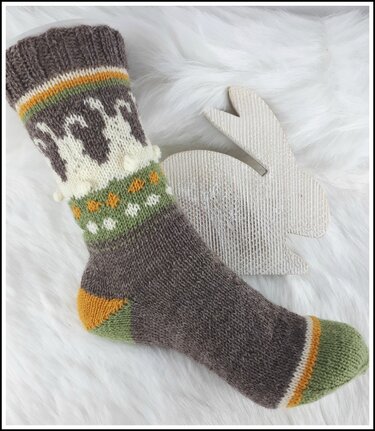 Braune gestrickte Socke mit weißen Hasenmotiven, grünen Punkten und orangefarbenen Akzenten auf weißem Fellhintergrund neben einem Holzhasen.