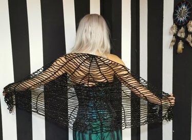 Rückansicht einer Person, die ein schwarzes, gehäkeltes Dreieckstuch im Spinnennetzmuster vor einem schwarz-weiß gestreiften Hintergrund trägt.