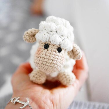 Close-up of a small crocheted amigurumi sheep held in a palm, showing bobble stitch texture and details.