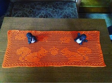 Orangefarbener gehäkelter Tischläufer im Filetmuster mit angedeutetem Tiermotiv auf Holztisch, zwei kleine schwarze Figuren