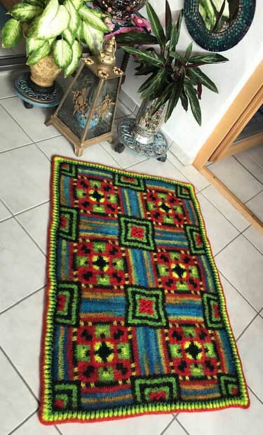 Handgehäkelter bunter Teppich mit geometrischem Quadrat- und Streifenmuster auf hellem Fliesenboden.