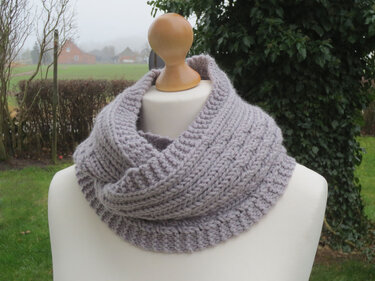 Hellgrauer gestrickter Loop-Schal mit Rippen- und Strukturmuster auf einer weißen Schaufensterpuppe vor unscharfem Außenhintergrund.