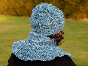 Rückenansicht einer Person mit hellblauer, gestrickter Zopfmuster-Mütze und passendem Halswärmer vor unscharfem Grün