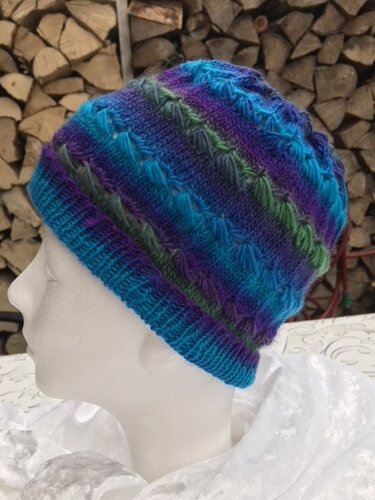 Gestrickte mehrfarbige Mütze in Blau, Grün und Lila mit Zopfmuster und Rippbündchen auf einer weißen Schaufensterpuppe vor gestapeltem Holz
