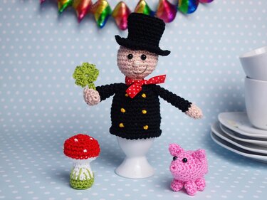 Gehäkelter Schornsteinfeger-Eierwärmer mit Kleeblatt, dazu Fliegenpilz und Glücksschwein vor gepunktetem Hintergrund.