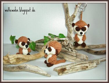 Drei gehäkelte Amigurumi-Erdmännchen in Braun und Beige, eines mit Schlüsselring, arrangiert auf Treibholz mit grünen Blättern