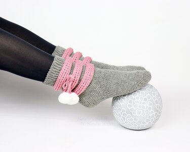 Grey crocheted socks with pink ribbed cuffs and white pom-poms worn over dark tights.