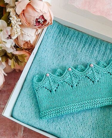 Türkisfarbenes gestricktes KronenStirnband mit Perlen auf hellblauer Strickdecke in weißer Geschenkbox, daneben rosa Blumen