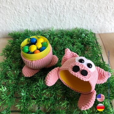 Two pink crocheted pig container covers on artificial grass, one holding colorful eggs.