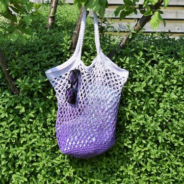 Gehäkelter Netzbeutel in Lila-Ombré hängt an Ast mit Sonnenbrille