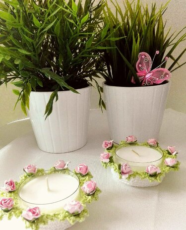 Nahaufnahme zweier Teelichthalter mit gehäkelten Rosenkränzen, arrangiert mit grünen Pflanzen und einem rosa Schmetterling