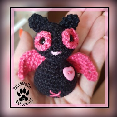 Gehäkeltes Mini-Fledermaus-Amigurumi in Schwarz mit pinken Flügeln, applizierten Augenringen und Herzknopf, in einer Hand gehalten