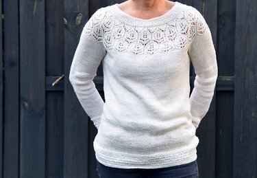 Heller gestrickter Damenpullover mit filigraner blattähnlicher Lochmuster-Passe am Rundhalsausschnitt, getragen vor dunklem Holzzaun.