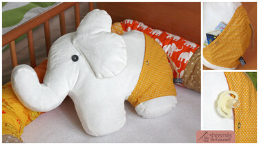 Weißes Elefanten-Plüschtier mit gelber, gepunkteter Stofftasche in einem Babybett mit orangefarbener Bettschlange