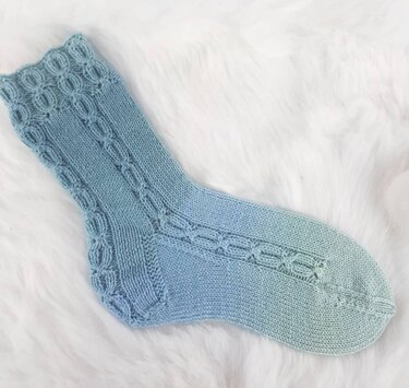 Gestrickte Socke in blaugrünem Farbverlauf mit Lochmuster auf weißem, flauschigem Untergrund