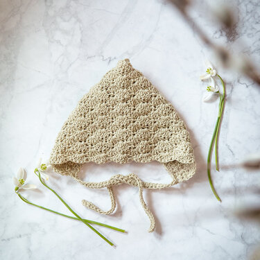 Hellbeige gehäkelte Babymütze mit Muschelmuster und Bindebändern auf hellem Marmor, dekoriert mit Schneeglöckchen.
