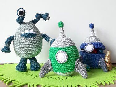 Drei gehäkelte Alien- und Raketenfiguren in Blau- und Grüntönen auf grüner Filzmatte
