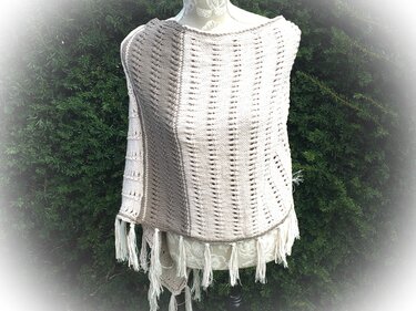 Beiger gestrickter Poncho mit Lochmuster und Fransen auf Schneiderpuppe vor grünen Hecken
