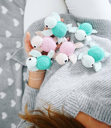 Hands holding small crocheted amigurumi turtles in pastel teal, pink, and cream.