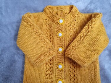 Gelbe gestrickte Babyjacke mit Blumenknöpfen und Ajourmuster auf grauem Hintergrund.