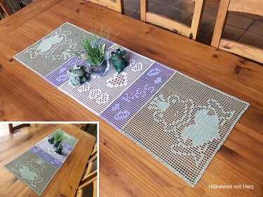 Gehäkelter Tischläufer mit Herz- und Blockmustern in Pastellgrün, Weiß und Lila auf einem Holztisch, dekoriert mit Froschfiguren und einer kleinen Pflanze.