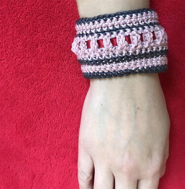 Nahaufnahme einer Hand mit breiter gehäkelter Armstulpe in Rosa und Dunkelgrau, rotes Band sichtbar, roter Hintergrund