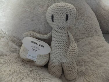 Hellbeige gehäkelte Amigurumi-Puppe mit gestickten Augen neben einem Garnknäuel auf flauschigem Untergrund