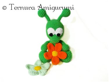 Green crocheted amigurumi with antennae holding an orange crochet flower, mint flower beside it