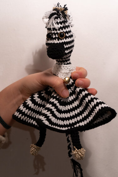 Schwarz-weiß gestreiftes, gehäkeltes Zebra-Schmusetuch mit Kopf, Glöckchen und Quasten, gehalten von einer Hand