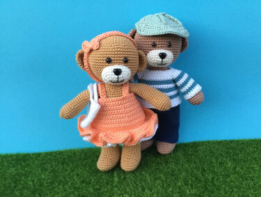 Zwei gehäkelte Amigurumi-Teddybären: einer im pfirsichfarbenen Rüschkleid mit Stirnband, der andere im gestreiften Pullover und Schirmmütze, vor blauem Hintergrund auf grünem Untergrund