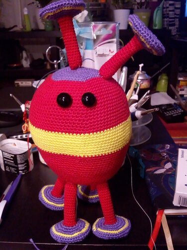 Rotes gehäkeltes Amigurumi mit gelben Streifen, lila Details und schwarzen Augen