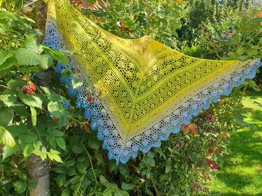 Gehäkeltes Dreieckstuch mit Gelb‑Blau‑Farbverlauf und Lochspitze, an einem Ast im Garten aufgehängt.