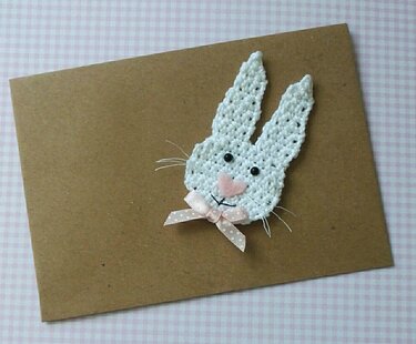 Weißer Häkel-Hasenkopf mit langen Ohren, rosa Filznase und gepunkteter Schleife auf brauner Karte