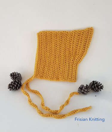 Mustard-yellow crocheted baby bonnet with long ties laid flat on a white background beside pine cones